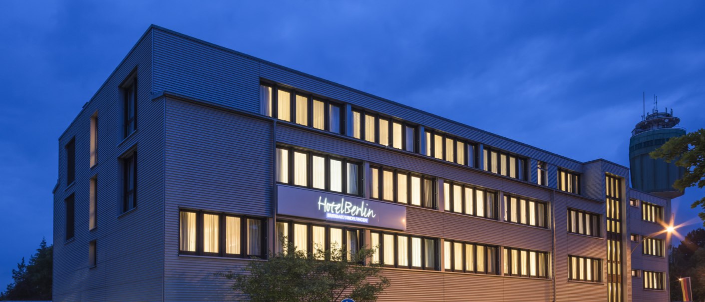 A modern hotel building at night, with illuminated windows and a blue sky. Cars on the street, leaving trails of light. The building bears the sign &ldquo;Hotel Berlin.&rdquo;, &copy; Hotel Berlin aZIS Hotel BetriebsGmbH