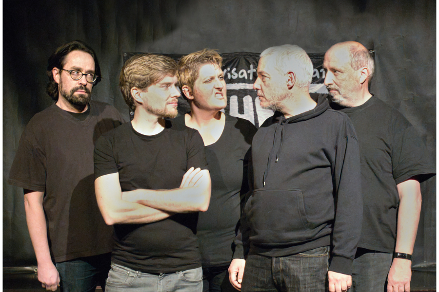 F&uuml;nf Personen in schwarzer Kleidung stehen auf einer B&uuml;hne und schauen sich an. Hintergrundbanner mit Schriftzug., &copy; Fotocredit Improvisationstheater Ohne Gew&auml;hr