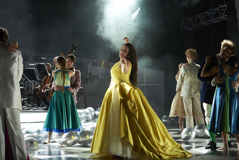 Menschen in eleganter Kleidung tanzen auf einer Bühne. Eine Frau im gelben Kleid steht im Vordergrund. Musiker sind im Hintergrund zu sehen., © Württembergische Staatstheater Stuttgart Menschen in eleganter Kleidung tanzen auf einer Bühne. Eine Frau im gelben Kleid steht im Vordergrund. Musiker sind im Hintergrund zu sehen., © Württembergische Staatstheater Stuttgart