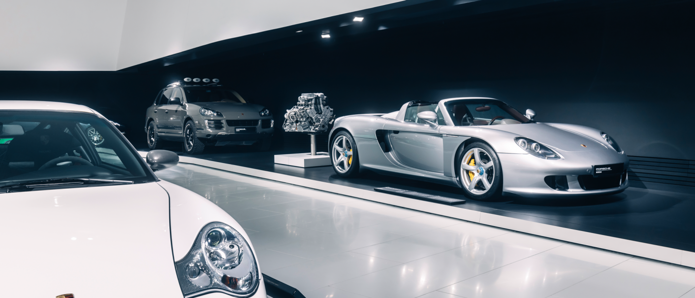 Three Porsche models in a modern showroom: a silver sports car, an SUV and a white car in the foreground. An engine is also on display., &copy; Porsche AG