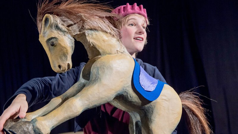 Eine Person mit rotem Hut präsentiert ein hölzernes Pferd mit blauer Decke. Der Hintergrund ist dunkel, was die Szene hervorhebt., © FITZ Das Theater animierter Formen