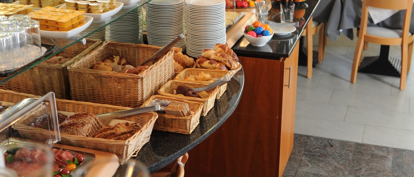 Ein Frühstücksbuffet mit Gebäck, Müsli, Obst und Aufschnitt in einem hellen Speiseraum mit gedeckten Tischen., © TOMAS