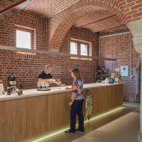 Eine Theke in einer Weinkellerei mit Backsteinwänden. Ein Mann schenkt einer Frau Wein ein. Weinflaschen sind auf der Theke ausgestellt., © Collegium Wirtemberg Eine Theke in einer Weinkellerei mit Backsteinwänden. Ein Mann schenkt einer Frau Wein ein. Weinflaschen sind auf der Theke ausgestellt., © Collegium Wirtemberg