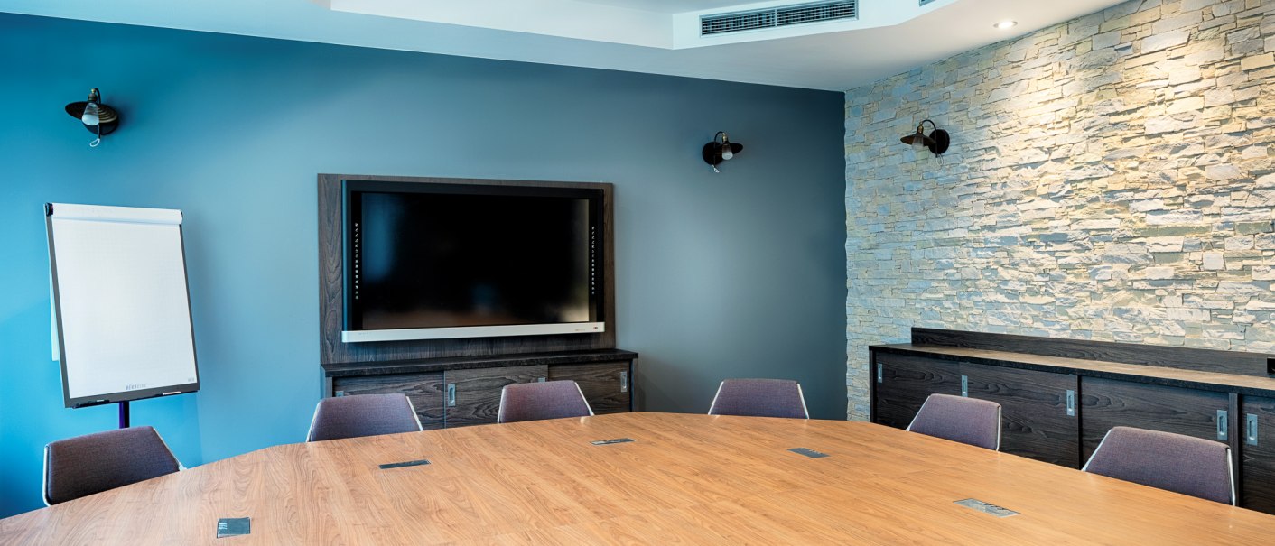 Moderner Tagungsraum mit gro&szlig;em Holztisch, St&uuml;hlen, einem Bildschirm an der Wand und einem Flipchart. Die W&auml;nde sind blau und mit Steinverkleidung., &copy; Premier Inn Germany