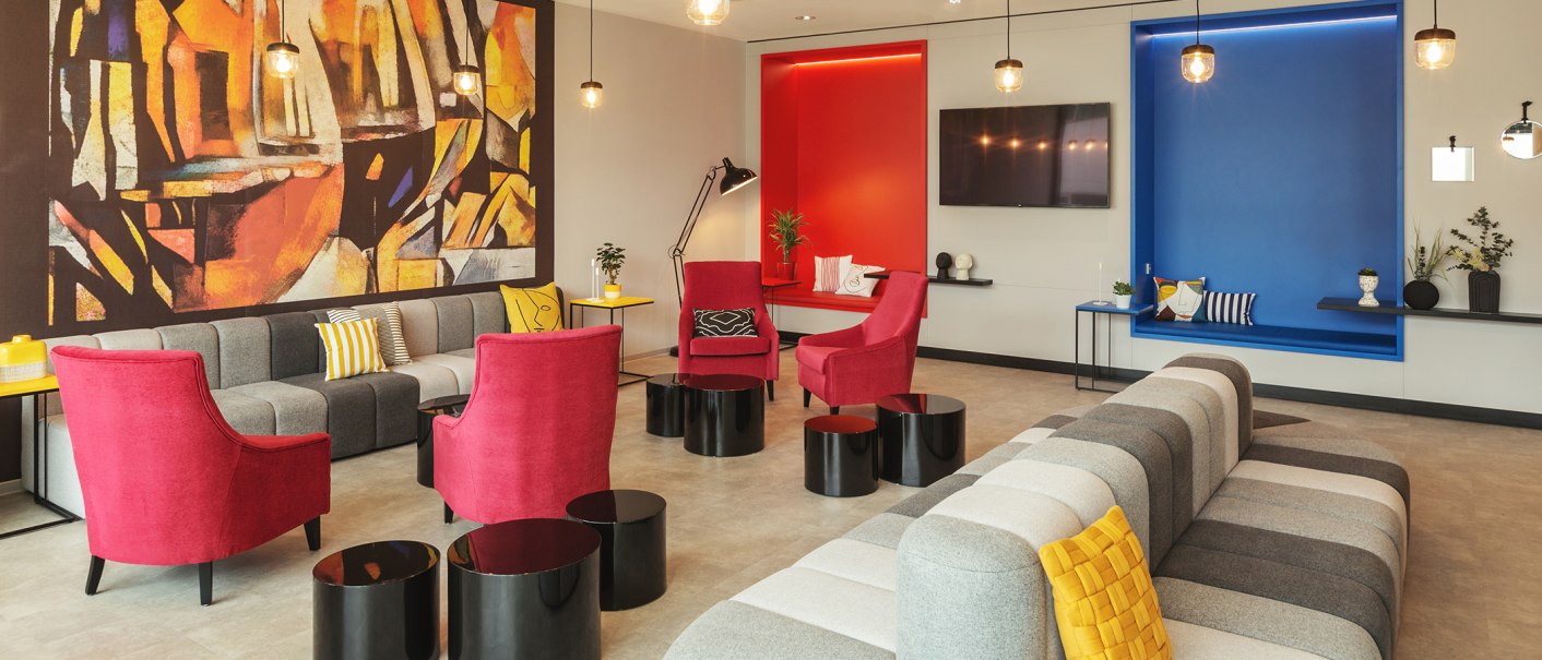 A modern lobby with gray and red seating, colorful cushions, an abstract mural, and colorful alcoves in red and blue., © the niu Form - Novum Hospitality A modern lobby with gray and red seating, colorful cushions, an abstract mural, and colorful alcoves in red and blue., © the niu Form - Novum Hospitality