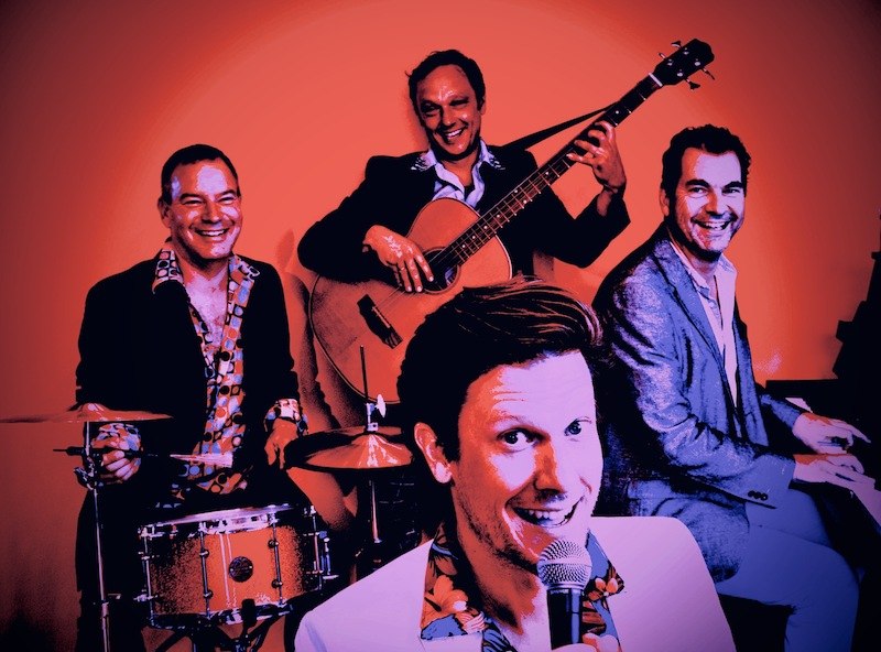 Vier Männer in Anzügen musizieren fröhlich. Einer singt ins Mikrofon, die anderen spielen Schlagzeug, Gitarre und Klavier. Der Hintergrund ist orange., © Rosenau Kultur e.V.
