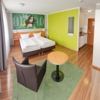 Modernes Superior Doppelzimmer mit zwei Betten, grünem Wandbild, Tisch, Stuhl und kleiner Küchenzeile auf Holzboden., © attimo Hotel Stuttgart