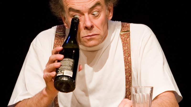 A man with a bald head and suspenders looks through a bottle. There is a glass on the table in front of him., &copy; Theaterhaus Stuttgart e.V.