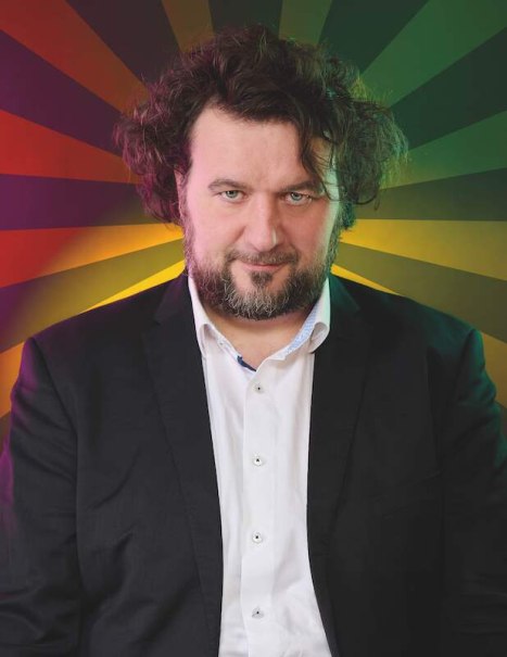 Ein Mann mit lockigem Haar und Bart trägt einen Anzug. Der Hintergrund ist bunt und strahlenförmig gestaltet., © Rosenau Kultur e.V. Ein Mann mit lockigem Haar und Bart trägt einen Anzug. Der Hintergrund ist bunt und strahlenförmig gestaltet., © Rosenau Kultur e.V.