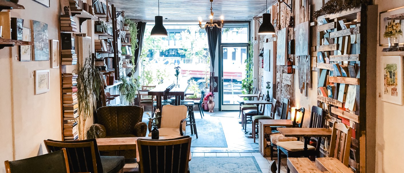 Ein gemütliches Café mit Holzmöbeln, Bücherregalen und Pflanzen. Große Fenster lassen viel Licht herein, und die Atmosphäre wirkt einladend und entspannt., © Café Che