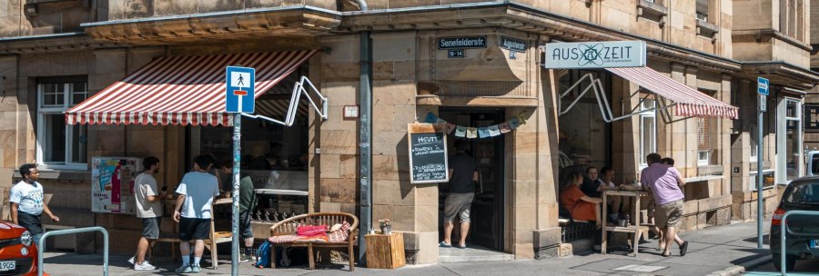 Caf&eacute;bar Auszeit on a street corner with a red awning. People are sitting outside, street signs and cars are visible., &copy; SMG, Sarah Schmid