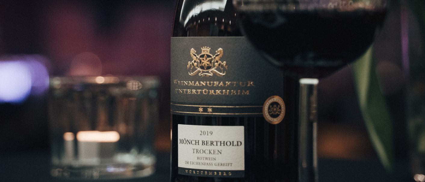 Nahaufnahme einer Weinflasche "Mönch Berthold" von 2019, trocken, mit einem Glas Rotwein daneben. Elegantes Etikett mit goldener Schrift., © Weinmanufaktur Untertürkheim Nahaufnahme einer Weinflasche "Mönch Berthold" von 2019, trocken, mit einem Glas Rotwein daneben. Elegantes Etikett mit goldener Schrift., © Weinmanufaktur Untertürkheim