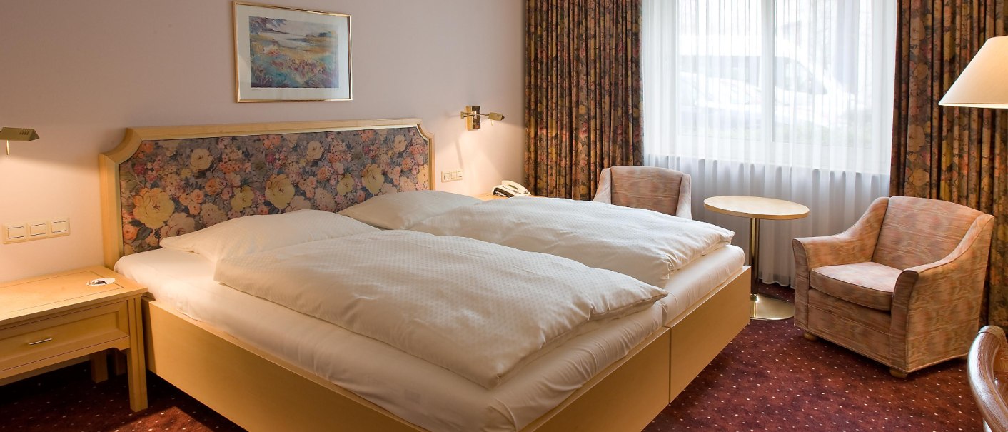 Comfortable hotel room with double bed, floral headboard, two armchairs, small table and window with curtains., © Erikson Hotel