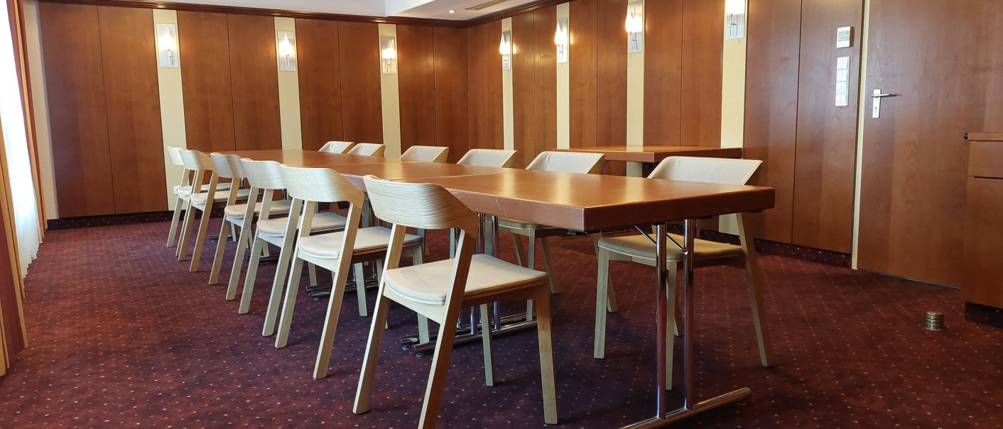 An empty meeting room with wooden tables and chairs on a red carpeted floor. The walls are wood-paneled and there is subtle lighting., &copy; Erikson Hotel