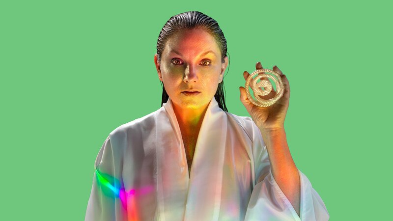 Person in weißem Gewand hält ein spiralförmiges Objekt vor grünem Hintergrund. Regenbogenfarben reflektieren auf dem Gewand., © Württembergische Staatstheater Stuttgart