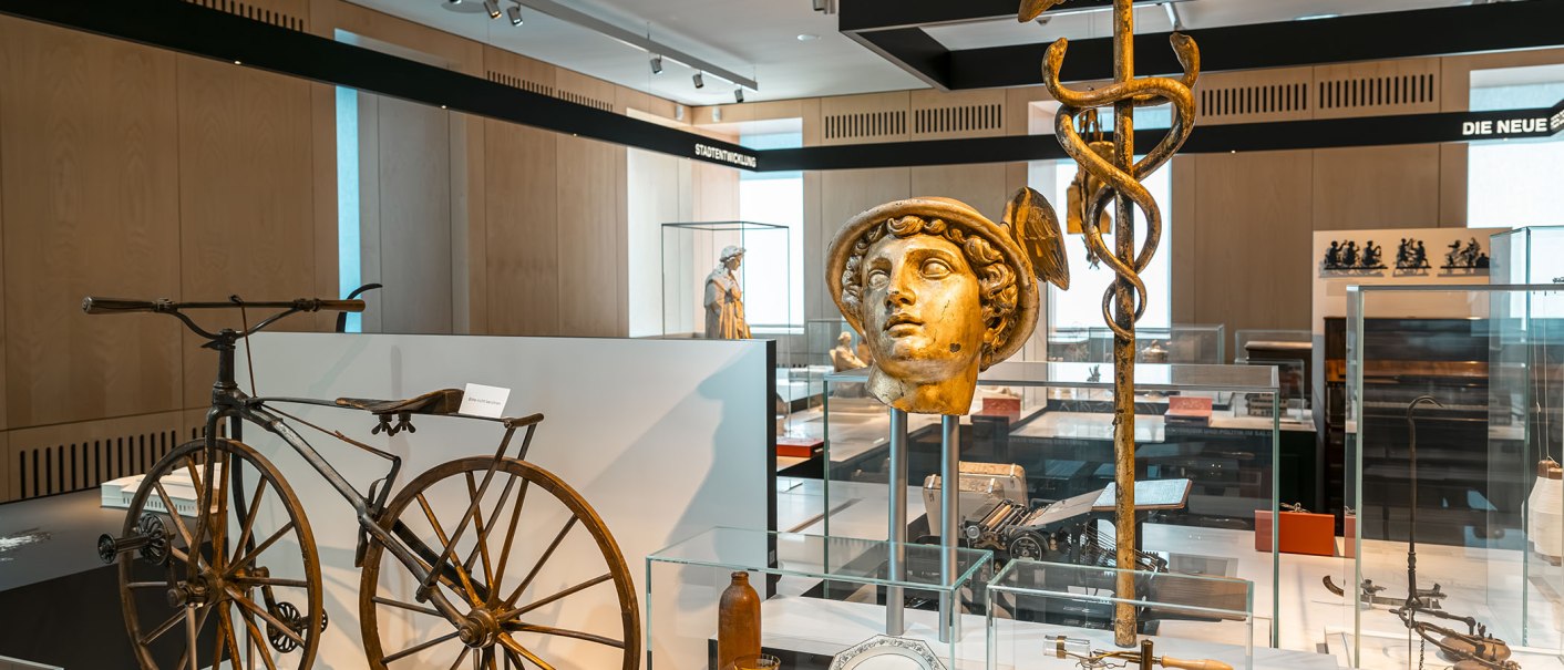 Ein Ausstellungsraum im StadtPalais zeigt antike Objekte, darunter ein altes Fahrrad, eine goldene Büste und ein Hermesstab. Moderne Beleuchtung und Vitrinen., © Stuttgart Marketing GmbH, Sarah Schmid
