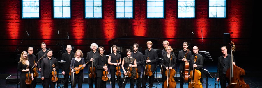 Das Stuttgarter Kammerorchester posiert auf einer Bühne vor einer roten Backsteinwand mit großen Fenstern. Die Musiker halten ihre Instrumente., © Reiner Pfisterer Das Stuttgarter Kammerorchester posiert auf einer Bühne vor einer roten Backsteinwand mit großen Fenstern. Die Musiker halten ihre Instrumente., © Reiner Pfisterer