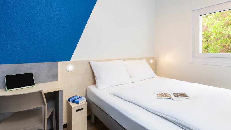 Ein schlichtes Hotelzimmer mit Doppelbett, blau-wei&szlig;er Wand, kleinem Schreibtisch und Fenster mit Blick ins Gr&uuml;ne., &copy; DQuadrat Living GmbH