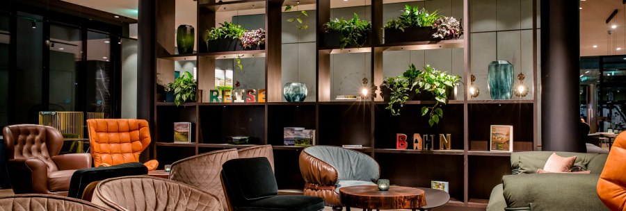 Eine stilvolle Lobby mit bequemen Sesseln, Pflanzen und dekorativen Regalen. Die Atmosph&auml;re ist einladend und modern., &copy; Motel One - Stuttgart Hauptbahnhof