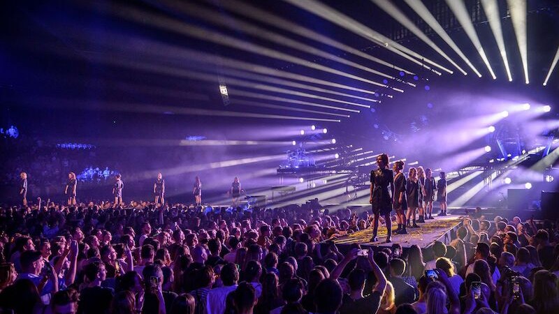 Konzertszene mit lila Lichtern, Models auf einem Laufsteg und einer großen Menschenmenge. Strahlenförmige Beleuchtung erzeugt eine dynamische Atmosphäre., © Fratelli BV