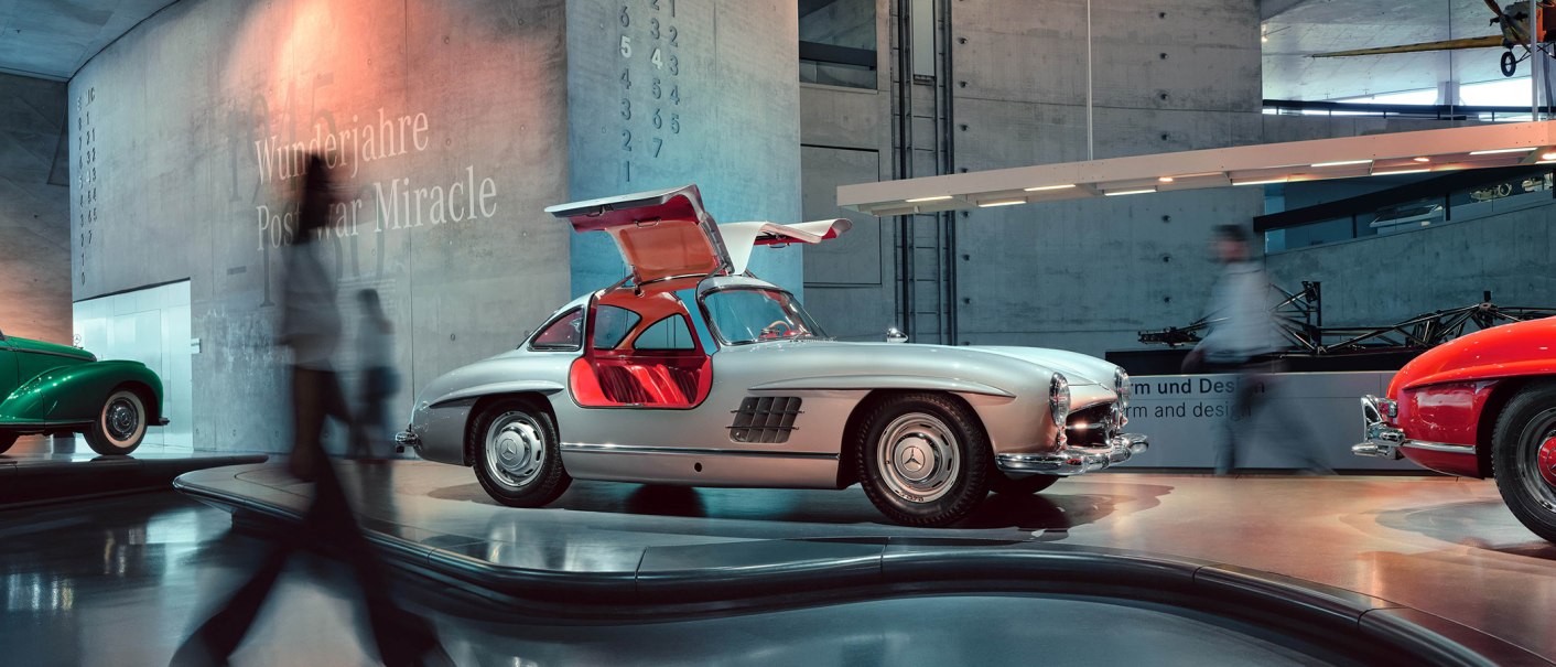 Silver vintage car with gullwing doors in the Mercedes-Benz Museum. People walk past, modern architecture in the background., © Mercedes-Benz Heritage GmbH Silver vintage car with gullwing doors in the Mercedes-Benz Museum. People walk past, modern architecture in the background., © Mercedes-Benz Heritage GmbH