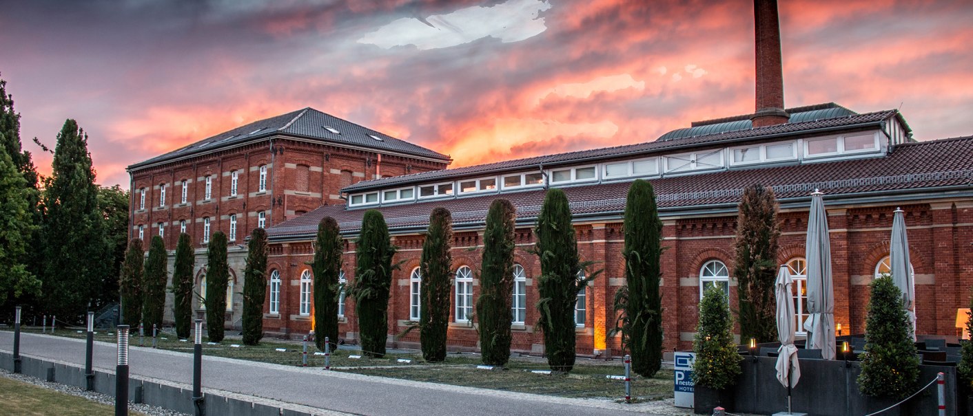 Historisches Backsteingebäude mit Bäumen und Schirmen im Vordergrund, bei dramatischem Sonnenuntergangshimmel., © nestor Hotel Ludwigsburg