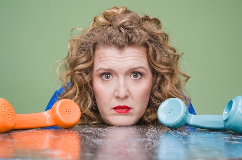 A woman with curly hair and red lipstick is lying on a table, looking out from between an orange and a blue telephone receiver., &copy; Rosenau Kultur e.V.