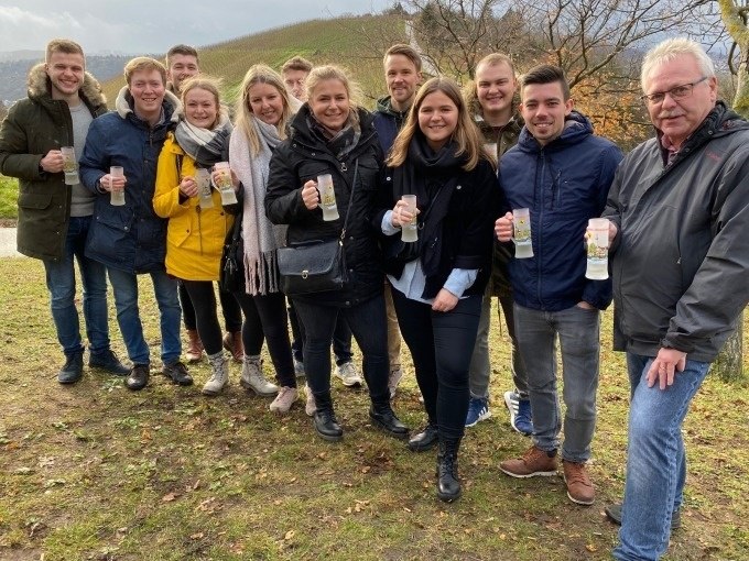 Eine Gruppe von Menschen steht lächelnd in einem Park und hält Glühweintassen. Im Hintergrund sind Bäume und Hügel zu sehen., © Weingenuss Stuttgart Eine Gruppe von Menschen steht lächelnd in einem Park und hält Glühweintassen. Im Hintergrund sind Bäume und Hügel zu sehen., © Weingenuss Stuttgart