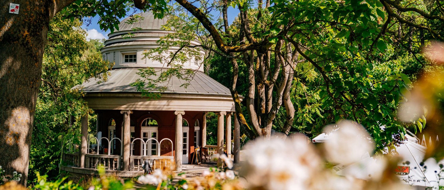 Ein rundes Teehaus mit Säulen im Weißenburgpark, umgeben von grünen Bäumen und blühenden Blumen im Vordergrund., © Thomas Niedermüller Ein rundes Teehaus mit Säulen im Weißenburgpark, umgeben von grünen Bäumen und blühenden Blumen im Vordergrund., © Thomas Niedermüller