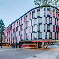 Modernes Hotelgeb&auml;ude mit bunter Fassade, Hilton Garden Inn Stuttgart Neckarpark. Vor dem Geb&auml;ude stehen Fahnenmasten, umgeben von B&auml;umen., &copy; Hilton Garden Inn Stuttgart Neckarpark