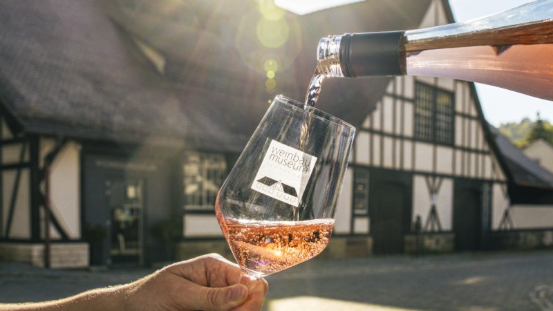 Roséwein wird vor dem Weinbaumuseum in ein Glas eingeschenkt, im Hintergrund ein Fachwerkgebäude im Sonnenlicht., © SMG, Sarah Schmid Roséwein wird vor dem Weinbaumuseum in ein Glas eingeschenkt, im Hintergrund ein Fachwerkgebäude im Sonnenlicht., © SMG, Sarah Schmid