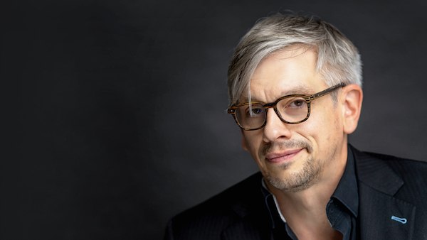 A man with glasses and gray hair smiles slightly against a dark background., © Renitenztheater Stuttgart e.V. A man with glasses and gray hair smiles slightly against a dark background., © Renitenztheater Stuttgart e.V.