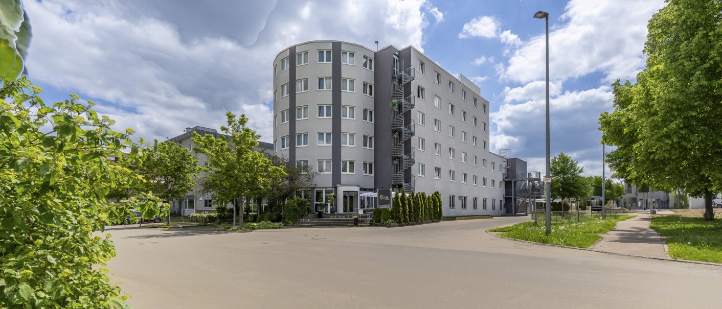 Modern, multi-story building with round corner, gray facade, surrounded by trees and sidewalk, under a cloudy sky., © Plaza INN Stuttgart-Filderstadt Modern, multi-story building with round corner, gray facade, surrounded by trees and sidewalk, under a cloudy sky., © Plaza INN Stuttgart-Filderstadt