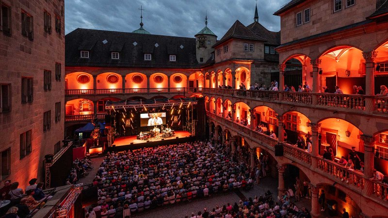 Open-air concert in the courtyard of a historic building, illuminated in red. Spectators sit on balconies and in the courtyard, stage with musicians., &copy; jazzopen stuttgart, Reiner Pfisterer