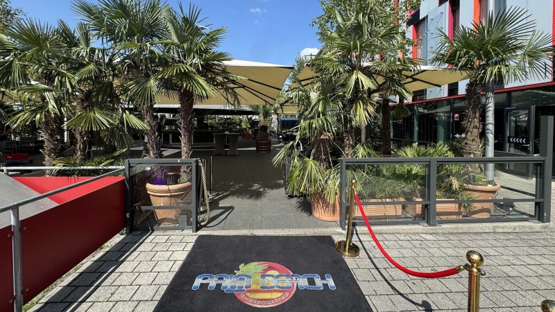Eingang zu einem Außenbereich mit Palmen, Sonnenschirmen und einem roten Teppich mit 'Palm Beach' Logo. Der Himmel ist blau., © Palm Beach Stuttgart GmbH Eingang zu einem Außenbereich mit Palmen, Sonnenschirmen und einem roten Teppich mit 'Palm Beach' Logo. Der Himmel ist blau., © Palm Beach Stuttgart GmbH