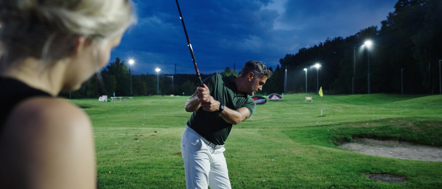 A man making a golf swing on a floodlit course at dusk, observed by a woman in the foreground., © GolfKultur Stuttgart A man making a golf swing on a floodlit course at dusk, observed by a woman in the foreground., © GolfKultur Stuttgart