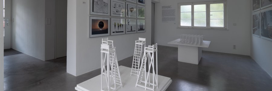 Heller Ausstellungsraum der Architekturgalerie mit gerahmten Arbeiten, Texttafel und weißen Architekturmodellen auf Podesten., © Thomas Fütterer Heller Ausstellungsraum der Architekturgalerie mit gerahmten Arbeiten, Texttafel und weißen Architekturmodellen auf Podesten., © Thomas Fütterer