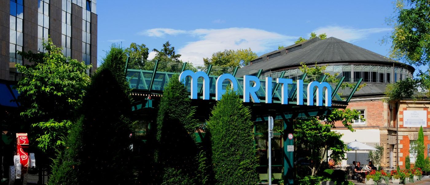 Exterior view of the Maritim Hotel with striking glass roof and surrounded by trees and plants. A path runs alongside the building., © Maritim Hotelgesellschaft mbH Exterior view of the Maritim Hotel with striking glass roof and surrounded by trees and plants. A path runs alongside the building., © Maritim Hotelgesellschaft mbH