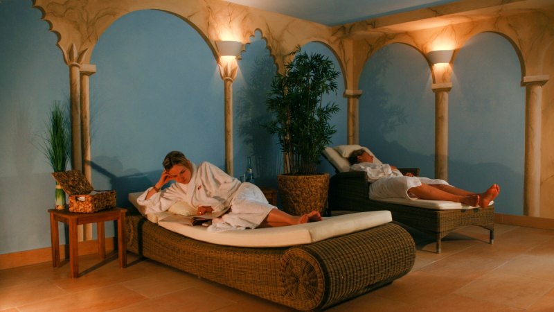 Zwei Personen in weißen Bademänteln entspannen auf Liegen in einem stilvollen Spa-Raum mit blauen Wänden und dekorativen Säulen., © TOMAS Zwei Personen in weißen Bademänteln entspannen auf Liegen in einem stilvollen Spa-Raum mit blauen Wänden und dekorativen Säulen., © TOMAS