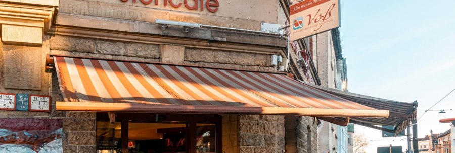 A stand-up café with a striped awning and the sign 'Voß' on the façade. The sun shines on the building., © SMG, Sarah Schmid A stand-up café with a striped awning and the sign 'Voß' on the façade. The sun shines on the building., © SMG, Sarah Schmid