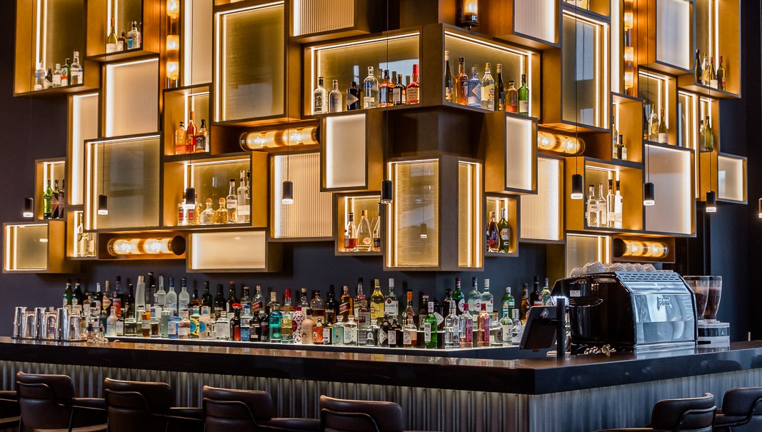 Eine stilvolle Bar mit beleuchteten Regalen voller Flaschen, modernen Barhockern und einer eleganten Theke., © TOMAS