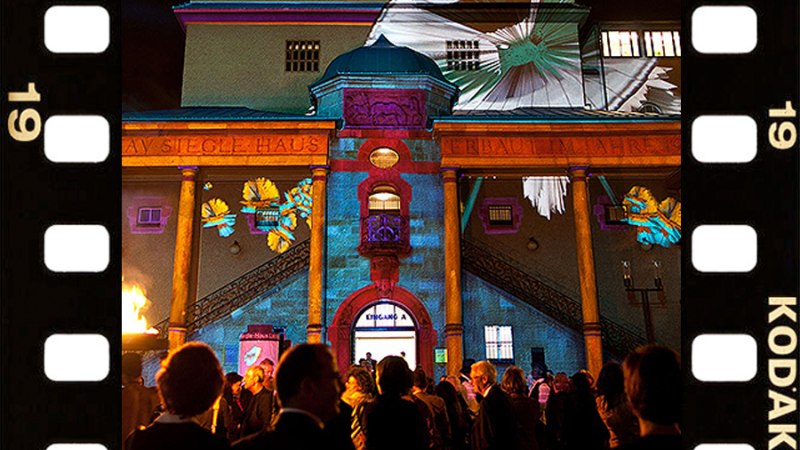 Das Gustav-Siegle-Haus ist nachts bunt beleuchtet. Eine Menschenmenge steht davor. Links und rechts sind Filmstreifen zu sehen., © Stuttgarter Philharmoniker Das Gustav-Siegle-Haus ist nachts bunt beleuchtet. Eine Menschenmenge steht davor. Links und rechts sind Filmstreifen zu sehen., © Stuttgarter Philharmoniker