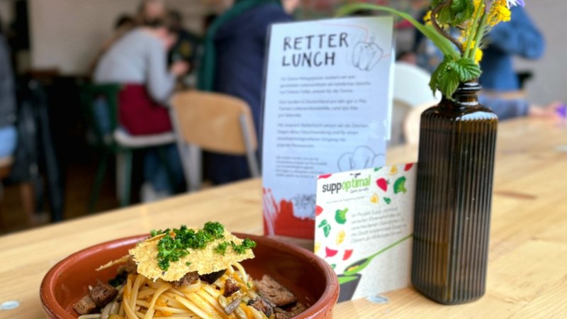 Ein Teller mit Pasta und Kräutern steht auf einem Tisch in einem Café. Daneben eine Vase mit Blumen und ein Schild mit der Aufschrift 'Retter Lunch'., © Foto©Labyrinth Ein Teller mit Pasta und Kräutern steht auf einem Tisch in einem Café. Daneben eine Vase mit Blumen und ein Schild mit der Aufschrift 'Retter Lunch'., © Foto©Labyrinth
