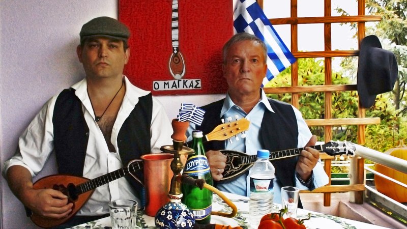 Zwei Männer in traditioneller Kleidung spielen griechische Instrumente an einem Tisch mit Getränken, Tomaten und einer griechischen Flagge im Hintergrund., © Fotocredit Theater La Lune/LEFTA rembetiko