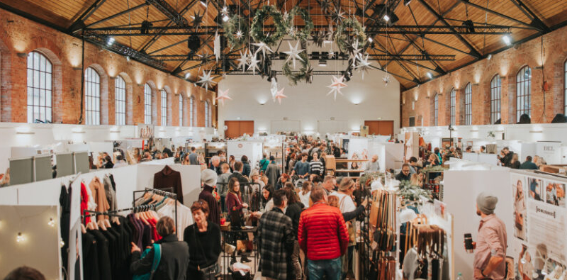 Große Halle mit Weihnachtsmarkt, dekoriert mit Sternen und Kränzen. Viele Menschen schlendern zwischen den Ständen mit Kleidung und Accessoires., © BLICKFANG Messe GmbH Große Halle mit Weihnachtsmarkt, dekoriert mit Sternen und Kränzen. Viele Menschen schlendern zwischen den Ständen mit Kleidung und Accessoires., © BLICKFANG Messe GmbH