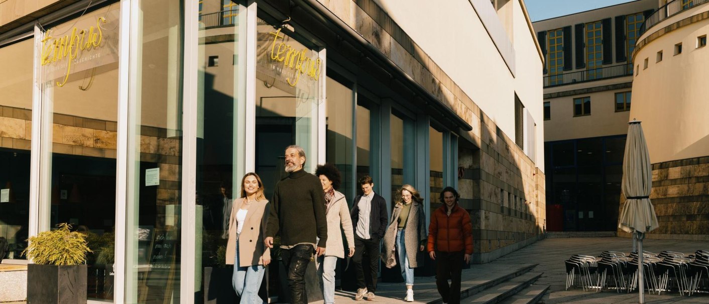 A group of people leave the Tempus Café & Bistro, a modern building with a glass façade. In the foreground are stacked chairs and a closed parasol., © Stuttgart-Marketing GmbH, Alwin Maigler A group of people leave the Tempus Café & Bistro, a modern building with a glass façade. In the foreground are stacked chairs and a closed parasol., © Stuttgart-Marketing GmbH, Alwin Maigler