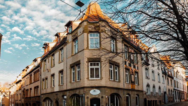 Eckgebäude mit roten Ziegeldächern, verziertem Mauerwerk und großen Fenstern. Ein Baum ohne Blätter steht daneben, blauer Himmel im Hintergrund., © Stuttgart-Marketing GmbH, Sarah Schmid