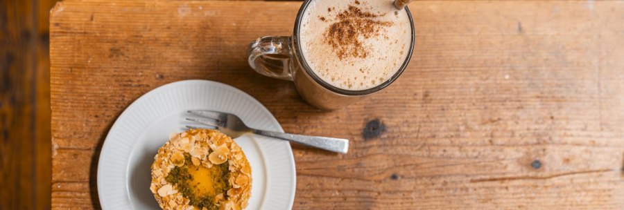 A plate of pastries garnished with almonds and a cup of coffee with milk foam on a rustic wooden table., © SMG, Sarah Schmid