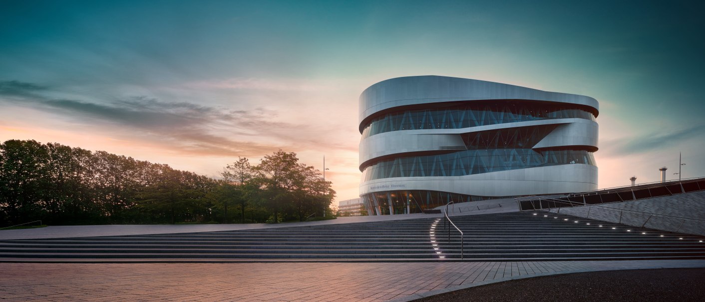 The Mercedes-Benz Museum in Stuttgart at sunset. Modern architecture with curved lines, surrounded by trees and illuminated steps., © Mercedes-Benz Heritage GmbH