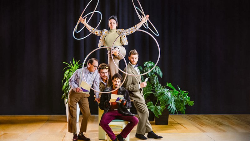 Eine Frau jongliert mit Reifen auf einem Stuhl, umgeben von vier M&auml;nnern, die konzentriert lesen. Pflanzen und ein dunkler Vorhang im Hintergrund., &copy; Theaterhaus Stuttgart e.V.